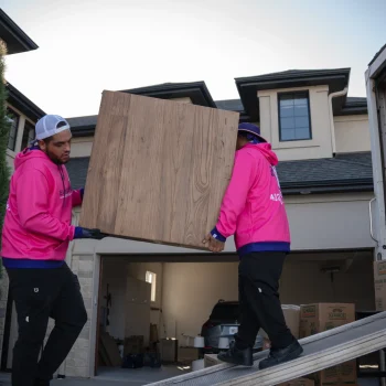 Two Easy as Pie movers in bright pink uniforms carrying a large wooden cabinet during a residential move in Omaha, showing their professional packing and long-distance moving service. Piano Movers Omaha: Safe Instrument Moving Experts